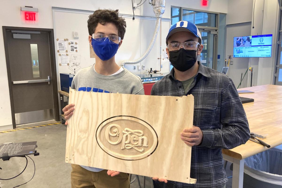 Two men holding an Open sign carved into wood in the Ruby Studio