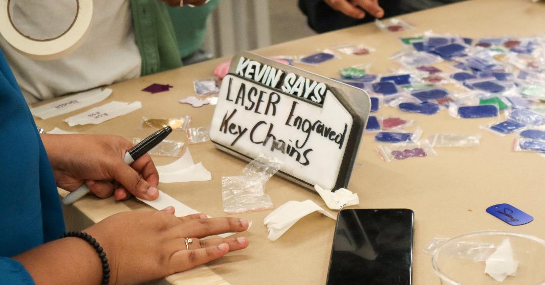 Image of keychains on a table with hands on the left-hand side holding a market and tape. Sign in the middle of the table says "Kevin says Laser Engraved Key Chains"
